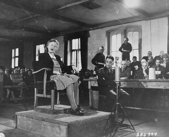 vrouw in jurk met donker jasje zit in stoel op een klein podium, met achter en naast haar mensen die luisteren