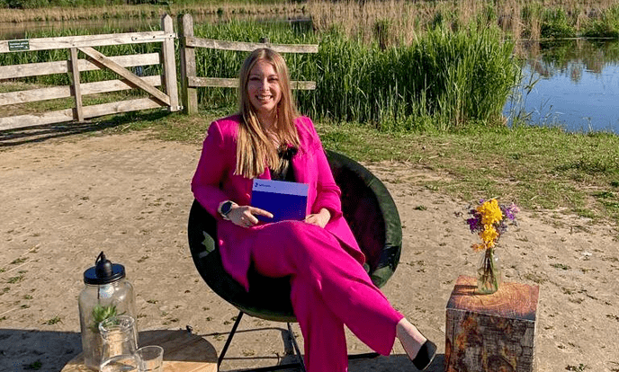 Anieke Kranenburg draagt een roze pak en zit op een stoel voor een meer