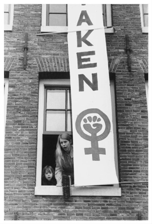 lang laken hangt uit raam van een huis met daarop een gedeelte van het woord staken en het feministisch symbool eronder